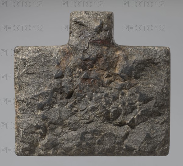 Nesichnum's sacrificial table; sacrificial formulas, vases, breads, cartouches, Nesichnum ca. 332–330 BC; ritual vessels iconographic; hieroglyphs; Ptolemaic Period, Egypt 332–30 BC; sacrificial table; height 5.5 cm, width 23 cm, depth 23 cm., A roughly rectangular stone slab with a small raised rectangular projection at the top, showing a coarse, uneven surface texture with pitted areas and angular indentations; the stone displays a mix of gray tones with darker charcoal patches and subtle brownish and rusty ochre areas, and the background behind the stone is a smooth neutral gray.