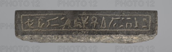 Nesichnum's sacrificial table; sacrificial formulas, vases, breads, cartouches, Nesichnum ca. 332–330 BC; ritual vessels iconographic; hieroglyphs; Ptolemaic Period, Egypt 332–30 BC; sacrificial table; height 5.5 cm, width 23 cm, depth 23 cm;, A rectangular stone slab with a rough, irregular lower edge rests against a neutral gray background; the top face is darker with a slightly textured surface and shows the impression of a framed rectangular panel across its length, while the lower exposed edge reveals lighter gray and beige mineral variations.