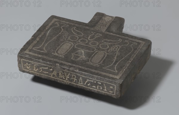 Nesichnum's sacrificial table; sacrificial formulas, vases, breads, cartouches, Nesichnum ca. 332–330 BC; ritual vessels iconographic; hieroglyphs; Ptolemaic Period, Egypt 332–30 BC; sacrificial table; height 5.5 cm, width 23 cm, depth 23 cm, A dark gray rectangular stone slab with a short protruding tab rests on a light gray surface, showing lighter carved outlines on its top face that form a symmetrical arrangement of abstract shapes and vessel-like forms; the slab's edges are worn and reveal a few rough brownish spots, and the object casts a soft shadow to the right.