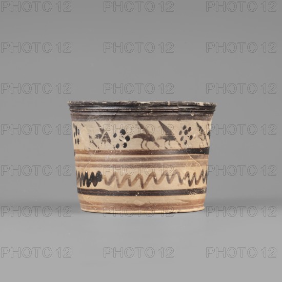 Pyxis with a duck motif. Vessel of yellowish clay with dark brown matte paint and brown varnish; flat bottom, walls slightly flaring to a profiled rim. Banded decoration: upper band shows a row of birds ducks walking to the right in silhouette with heads lowered and wings raised, with dotted rosettes between pairs; lower band enclosed in two wide dark bands consisting of two stripes of watered varnish and a zigzag line. Greek, Boeotia; early 7th century BC. Pyxis container. Clay color: 10 YR 7/2–7/4; varnish color: 2.5 YR 2.5/1; red paint: 2.5 YR 6/3. Dimensions: depth 5.5 cm; diameter 7.3 cm; wall thickness 0.4 cm; height 6.5 cm; width 8.8 cm; weight 0.098 kg., A short cylindrical ceramic vessel sits centered against a plain gray background, showing a beige base color with bands of darker brown and black decoration. The rim is dark brown and slightly worn, and just below it are painted motifs including abstract bird-like shapes and clustered round dots in black. Midsection bands alter...