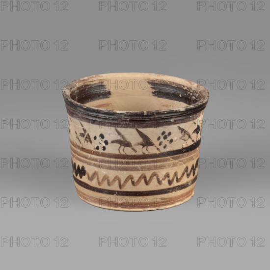 Pyxis with a duck motif. Ceramic vessel pyxis of yellowish clay with dark brown matte paint and brown varnish. Flat bottom, walls slightly flaring toward a profiled rim. Banded decoration: upper band shows a row of birds ducks walking to the right in silhouette with lowered heads and raised wings; dotted rosettes between pairs of birds. Below, decoration enclosed in two wide dark bands consisting of two stripes of watered varnish and a zigzag line. Clay color: 10 YR 7/2–10 YR 7/4; varnish color: 2.5 YR 2.5/1 black; red paint: 2.5 YR 6/3. Early 7th century BC, Boeotia. Function: container. Dimensions: depth 5.5 cm; diameter 7.3 cm; wall thickness 0.4 cm; height 6.5 cm; width 8.8 cm; weight 0.098 kg., A cylindrical ceramic vessel sits on a flat surface against a neutral gray background, showing an open top and a rounded rim with worn dark brown areas. The vessel's exterior is primarily beige with decorative bands and patterns in dark brown and lighter brown tones; a band near the top fea...