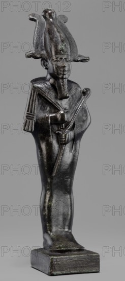 Figurine of the god Osiris, Osiris mythology; statuette of a standing Osiris wrapped in a long, tight-fitting robe forming a characteristic rounding at the back of the shoulders. On the head is an atef crown with horns and a uraeus. The feet rest on a rectangular hollow base with a tenon for seating on the base. The four sides of the base are covered with a hieroglyphic inscription in two lines. 664–525 BC; Late Period, 26th Dynasty, Egypt. Figurine; height 31.5 cm, width 9.4 cm., A dark metallic small statue of a standing human figure with a tall ornate headdress and a false beard, shown against a plain gray background; the figure wears a detailed broad collar and holds a curved staff across its body, with hands clasping the staff; the surface shows a glossy dark brown to black patina with areas of greenish corrosion and lighter worn highlights; the figure stands on a rectangular base that shares the same dark metallic color and slightly textured surface.