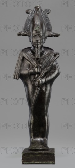 Figurine of the god Osiris, Statuette of a standing Osiris wrapped in a long, tight-fitting robe forming a characteristic rounding at the back of the shoulders. On the head is an atef crown with horns and a uraeus. The feet rest on a rectangular hollow base with a tenon for seating on the base. The four sides of the base are covered with a hieroglyphic inscription in two lines. Osiris mythology; atef crown; uraeus; Egyptian deities; mummy-shaped figurines; Egyptian art; Egyptian sculpture. Date: 664–525 BC, Late Period, 26th Dynasty, Egypt. Object type: figurine. Height: 31.5 cm; Width: 9.4 cm., A dark metallic statue of a human figure stands on a rectangular base against a gray background, the figure wearing a tall headdress with curved flaring elements and a central raised ridge, and a false beard extending from the chin; the figure's eyes, nose, and lips are visible with a straight facial expression, and there are textured details on the collar area and headdress; the figure's arms ...