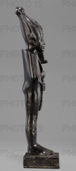 Figurine of the god Osiris, Statuette of a standing Osiris wrapped in a long, tight-fitting robe forming a characteristic rounding at the back of the shoulders. On the head is an atef crown with horns and a uraeus. The feet rest on a rectangular hollow base with a tenon for seating on the base. The four sides of the base are covered with a hieroglyphic inscription in two lines. Osiris mythology; Egyptian deities; mummy-shaped figurine; Egyptian art; Egyptian sculpture. 664–525 BC; Late Period, 26th Dynasty, Egypt. Figurine; height 31.5 cm, width 9.4 cm., A dark metallic standing figure shown in profile against a neutral gray background, with a tall rounded headdress extending backward, a stylized beard, and defined facial features; the figure has a smooth glossy surface with patches of lighter greenish oxidation, a broad flat back piece over the shoulders, arms held close to the body with hands clasping a vertical object, a slender torso and legs, and flat feet on a rectangular base th...