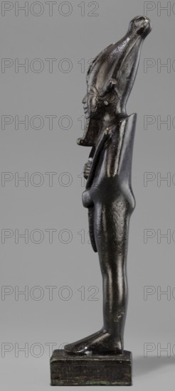 Figurine of the god Osiris. Statuette of a standing Osiris wrapped in a long, tight-fitting robe forming a characteristic rounding at the back of the shoulders. On the head is an atef crown with horns and a uraeus. The feet rest on a rectangular base, hollow inside, with a tenon for seating on the base. The four sides of the base are covered with a hieroglyphic inscription in two lines. Osiris mythology; Egyptian deities; mummy-shaped figurines; Egyptian art; Egyptian sculpture. 664–525 BC; Late Period, 26th Dynasty, Egypt. Statuette figurine. Height 31.5 cm; width 9.4 cm., A dark metallic figurine stands upright on a rectangular base against a plain gray background, showing a profile view of a humanlike figure with an elongated headdress that curves upward and backward. The figure's surface is glossy with reflections and areas of lighter sheen, and the overall color appears dark bronze with subtle greenish patina in places. The face has a pronounced nose and stylized features, and a s...