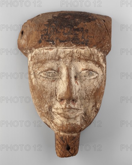 Sarcophagus mask of a deceased man. Creamy, elongated face, almond-shaped eyes, long straight nose. Small mouth with slightly upturned corners and prominent lips. Slightly arched eyebrows. Wig painted black on the head. Fragment of a braided beard under the chin. ca. 747–30 BC; Late Period–Ptolemaic period, Egypt., A carved wooden face with a rounded forehead and a blocky headdress, showing a weathered surface with a mix of light beige, warm brown, and dark brown tones; the facial features are lightly incised with closed or partially closed eyes, a defined nose, and a subtle mouth, all exhibiting worn paint and surface cracks; the headdress area is darker with rougher texture and patches of very dark brown, while the lower chin extends into a short rectangular wooden peg with visible grain and a hole, and the overall piece rests against a neutral light gray background.