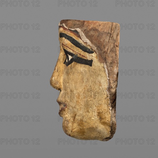 Woman's sarcophagus mask. Mask of wood covered with a layer of canvas with paint applied. The face is wide, painted yellow. Large almond-shaped eyes with pupils highlighted in black and arched eyebrows. Straight nose flattened at the tip. Mouth and chin protrusions marked in red, as is the outline of the eye sockets above the eyes. Face height: 15 cm. Sarcophagus fragments; masks, faces; grave goods. Mask from Egypt, 1st–2nd century; Roman period 4th century AD. Sarcophagus mask; height 15 cm, width 14.3 cm, depth 8 cm, weight 0.454 kg., A carved wooden profile of a human face oriented to the left with visible contours of a nose, lips, and chin, painted in a muted yellow-beige tone with areas of darker brown weathering; a painted eye area features bold black lines and a small white triangular highlight, with a band of reddish-brown and a thin yellow stripe above the eye; the top and back edges of the wood show natural brown wood grain and rough texture, and the background is a uniform ...