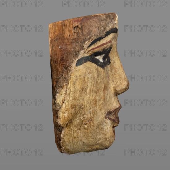 Woman's sarcophagus mask. Mask of wood covered with a layer of canvas to which paint is applied. The face is wide, painted with yellow paint. Large, almond-shaped eyes, the pupils highlighted with black paint, arched eyebrows. Straight nose, flattened at the tip. The mouth and the protrusions of the chin are marked in red, as is the outline of the eye sockets above the eyes. Face height: 15 cm. 1st–2nd century; Roman period 4th century AD. Egypt. Sarcophagus mask; height 15 cm, width 14.3 cm, depth 8 cm, weight 0.454 kg., A carved wooden profile of a human face shown against a gray background, with a textured surface and areas of worn paint; the face has visible eye makeup painted in black and dark blue with a small triangular white area, a band of dark reddish-brown above the eye, and lips painted in a muted red; the wood shows natural brown tones and a weathered creamy beige paint across the cheek and lower face, with rough patches and small cracks revealing darker brown grain.