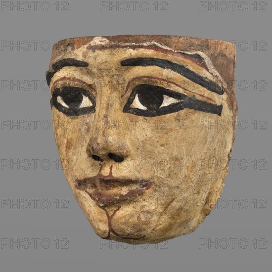 Woman's sarcophagus mask. Mask of wood covered with a layer of canvas to which paint is applied. The face is wide, painted with yellow paint. Large, almond-shaped eyes, the pupils highlighted with black paint, arched eyebrows. Straight nose, flattened at the tip. The mouth and the protrusions of the chin are marked in red, as is the outline of the eye sockets above the eyes. Face height: 15 cm. 1st–2nd century; Roman period 4th century AD, Egypt. Sarcophagus mask; height 15 cm, width 14.3 cm, depth 8 cm, weight 0.454 kg., A painted mask-like face carved from a light brown material with visible texture and wear, showing a few areas of darker brown and beige; the eyes are heavily outlined in black with white triangular shapes inside and dark pupils, and there are thick black painted eyebrows and a dark black stripe extending from one eye; there are faint reddish-brown painted lines above the eyes and around the mouth, with a darker reddish-brown used on the lips; the nose and lower face ...