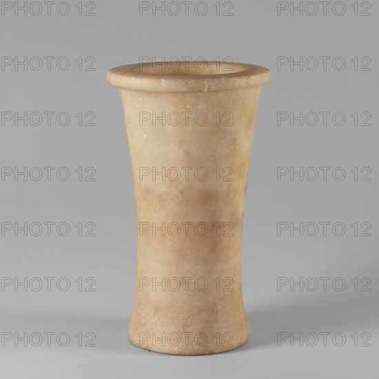 Bastet, stone vessel. The vessel walls taper gently to 1/3 of the height of the entire artifact, then begin to widen and end in a thick, flat rim. The bottom is slightly concave. Old Kingdom, ca. 2686–2181 BC; from Edfu. Height 26 cm, width 15.5 cm., A tall, cylindrical vase-like object with a flared rim and a slightly tapering body, shown against a plain background; the surface appears smooth and polished with subtle variations and marbling, and the visible colors include beige, creamy off-white, pale tan, and hints of light brown, with soft shadows around its base.