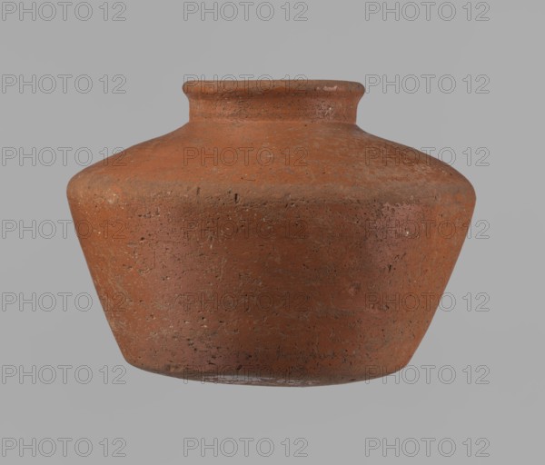Vessel, Egyptian pottery vessel clay, c. 2686–c. 2181 BC, Old Kingdom, Edfu; height 11 cm, rim diameter 6.3 cm, body diameter 14.7 cm, bottom diameter 10 cm., A single earthenware pot with a short neck and wide rounded body sits against a plain background, showing a coarse, textured surface with small pits and irregularities; the pot is primarily terracotta with variations of reddish-brown and hints of darker brown and muted orange, and the background appears as a neutral gray.