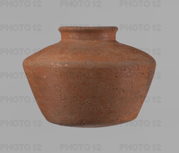 Vessel, Egyptian pottery, ceramic clay vessel, c. 2686–2181 BC Old Kingdom, from Edfu; height 11 cm, rim diameter 6.3 cm, body diameter 14.7 cm, bottom diameter 10 cm., A rounded, wide clay pot with a short narrow neck and flared rim sits against a plain background; the pot surface appears textured and slightly weathered with small pits and rough patches, and its colors include shades of terracotta, reddish-brown, rust, and hints of darker brown, while the background is a muted gray.