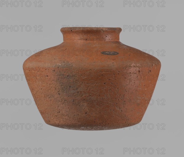 Vessel, Egyptian pottery vessel ceramic, clay, c. 2686–c. 2181 BC, Old Kingdom; excavated at Edfu. Vessel: height 11 cm; rim diameter 6.3 cm; body diameter 14.7 cm; bottom diameter 10 cm., A single rounded clay pot with a short neck and flared rim sits against a plain background; the pot has a rough, pitted surface texture with visible irregularities and wear, showing a primary reddish-brown color with areas of darker brown and small patches of lighter, almost pinkish tones, along with a few darker grayish spots and some subtle white residue near the base.