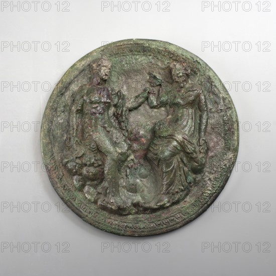 Mirror cover with an image of a couple. The bronze cover is decorated with a figurative scene of two people on the outside. The edge is also decorated with a braided ornament. The depiction includes a man and a woman facing each other, seated on rocks. The man's body is naked, with a chlamys draped over his left shoulder. The figure rests on a rock with his right hand, while his left touches the woman's right wrist. Dressed in a long chiton and himation, the woman also holds her left hand on a rock. The robe forms a few wide folds around the hips. A series of thick folds of fabric are also visible on the legs. Higher up, the fabric accentuates the woman's natural shape, while the folds are sparse and subtle. The depiction can be interpreted as a pair of lovers. Circular holes are carved in the man's right foot and the woman's left foot. Circular, c. 300 BC, Corinth; mirror; height 3 cm, diameter 14 cm., A round, relief-carved disc with a weathered, rough surface showing shades of green...