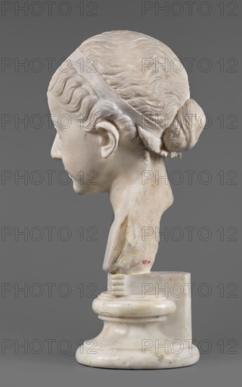 Portrait of a princess from the Julio-Claudian dynasty Julia?, unknown Roman workshop. The bust depicts a girl with a round, rather chubby face, turned slightly to the right; a rounded, elongated chin, and small, slightly pursed lips with a pronounced, wavy line of the upper lip. The almond-shaped, smooth eyes are rather shallowly set, with marked upper eyelids and barely visible eyebrows. The ears are carefully styled. The wavy hair is combed up and tied with a headband. At the back of the head, it is tied in a bun and a braid that falls down the middle of the head onto the smooth forehead. End of the 1st century BC–beginning of the 1st century AD, Roman Empire. Marble sculpture; height 25.7 cm, width 16 cm, depth 12.5 cm, weight 11.5 kg., A carved stone bust of a young person shown in profile against a plain gray background, rendered in off-white and pale beige tones with subtle darker beige and yellowish stains; the hair is styled back with visible wavy texture and gathered into a b...