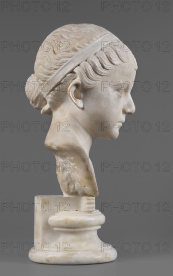 Portrait of a princess from the Julio-Claudian dynasty Julia?, unknown Roman workshop. The bust depicts a girl with a round, rather chubby face, turned slightly to the right; a rounded, elongated chin, and small, slightly pursed lips with a pronounced, wavy line of the upper lip. The almond-shaped, smooth eyes are rather shallowly set, with marked upper eyelids and barely visible eyebrows. The ears are carefully styled. The wavy hair is combed up and tied with a headband. At the back of the head, it is tied in a bun and a braid that falls down the middle of the head onto the smooth forehead. End of the 1st century BC–beginning of the 1st century AD, Roman Empire 27 BC–476. Sculpture. Height 25.7 cm, width 16 cm, depth 12.5 cm, weight 11.5 kg., The image shows a marble bust of a young person in profile against a solid gray background; the sculpture displays a smooth pale off-white surface with subtle beige and yellowish discolorations, carved wavy hair pulled back into a bun and held by...