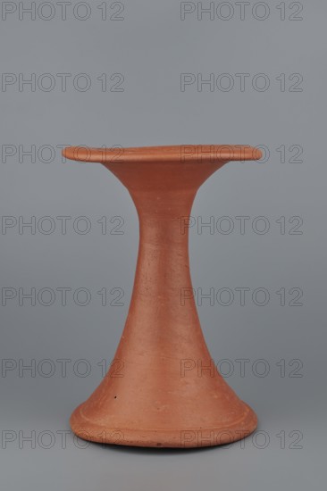 Bowl base. Base for a vessel with an almost equal diameter of both rims; both rims are irregular, with a wavy line. The center of the base is empty. The narrowest part of the base is approximately 2/3 of the height of the entire vessel. Made of Nile clay NB1. Surface leveled, covered with an engobe, then polished. Vessel color: red. Complete, very well preserved. c. 2494–c. 2181 BC; Old Kingdom, 5th–6th Dynasty, Edfu. Vessel; top diameter 17.5 cm, bottom diameter 18 cm, height 25.6 cm, weight 1.30 kg., A terracotta object shaped like a tall hourglass with a flared base and a flat, slightly rounded top is centered against a smooth gray background; the object has an earthy reddish-brown color with a matte, slightly rough surface showing small imperfections and subtle variations in tone, and the background displays a uniform cool gray.