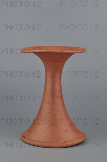 Bowl base, Base for a vessel with an almost equal diameter of both rims; both rims are irregular with a wavy line. The center of the base is empty. The narrowest part of the base is approximately 2/3 of the height of the entire vessel. Made of Nile clay NB1; surface leveled, covered with an engobe, then polished. Vessel color red. Complete vessel, very well preserved. c. 2494–c. 2181 BC; Old Kingdom, 5th–6th Dynasty, Edfu. Vessel; top diameter 17.5 cm, bottom diameter 18 cm, height 25.6 cm, weight 1.30 kg., A single terracotta-colored ceramic object with a flared base that tapers into a narrow stem and widens again into a flat, slightly convex top, set against a smooth gray background; the surface shows subtle texture variations and small imperfections in shades of reddish-brown and muted orange, with faint darker spots and lighter worn areas.