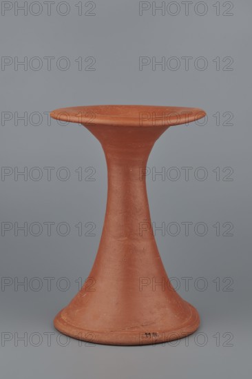 Bowl base, Base for a vessel with an almost equal diameter of both rims; both rims irregular with a wavy line. Center of the base is empty. The narrowest part of the base is approximately 2/3 of the height of the entire vessel. Vessel made of Nile clay NB1; surface leveled, covered with an engobe, then polished. Vessel color: red. Complete vessel, very well preserved. c. 2494–c. 2181 BC; Old Kingdom, 5th–6th Dynasty, Edfu. Vessel; top diameter 17.5 cm, bottom diameter 18 cm, height 25.6 cm, weight 1.30 kg., A terracotta-colored, hourglass-shaped ceramic object with a wide flared top and a broad rounded base set against a smooth gray background; the surface shows slight texture and small imperfections in the clay, with subtle variations of reddish-brown and orange tones and hints of darker speckles.