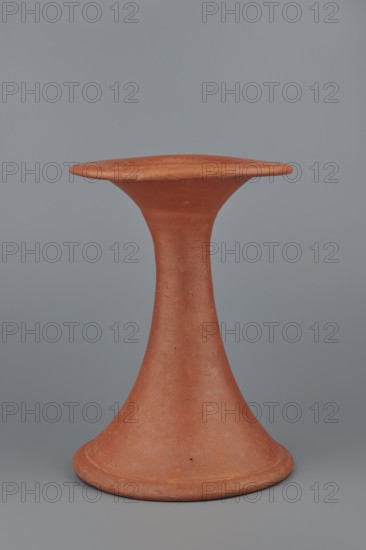 Bowl base. Base for a vessel with an almost equal diameter of both rims. Both rims are irregular, with a wavy line. The center of the base is empty. The narrowest part of the base is approximately 2/3 of the height of the entire vessel. Vessel made of Nile clay NB1. The surface is leveled and covered with an engobe, then polished. Vessel color: red. Complete vessel, very well preserved. c. 2494–c. 2181 BC; Old Kingdom, 5th–6th Dynasty, Edfu findplace. Vessel; diameter top 17.5 cm, diameter bottom 18 cm, height 25.6 cm, weight 1.30 kg., A terracotta object with a smooth matte surface in shades of reddish-brown and orange, shaped like an hourglass with a flared circular base and a wide, flat top; the object shows subtle surface texture and small imperfections, and it stands against a muted gray background.