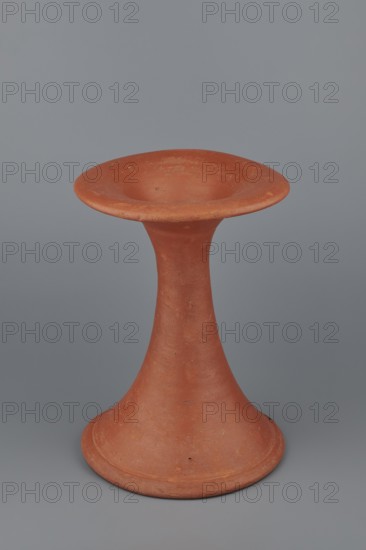 Bowl base. Base for a vessel with an almost equal diameter of both rims; both rims are irregular with a wavy line. The center of the base is empty. The narrowest part of the base is approximately 2/3 of the height of the entire vessel. Vessel made of Nile clay NB1. The surface is leveled and covered with an engobe, then polished. Vessel color: red. Complete vessel, very well preserved. c. 2494–c. 2181 BC; Old Kingdom, 5th–6th Dynasty, Edfu; vessel. Dimensions: diameter top 17.5 cm, diameter bottom 18 cm, height 25.6 cm; weight 1.30 kg., A terracotta-colored object with a flared circular base that tapers into a narrow neck and opens into a wide, shallow dish at the top, set against a solid gray background.