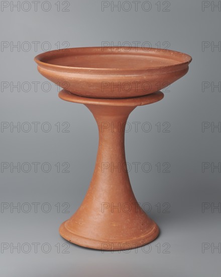 Bowl base. Base for a vessel with an almost equal diameter of both rims; both rims are irregular with a wavy line. The center of the base is empty. The narrowest part of the base is approximately 2/3 of the height of the entire vessel. Made of Nile clay NB1; surface leveled, covered with an engobe and then polished. Vessel color: red. Complete vessel, very well preserved. c. 2494–c. 2181 BC; Old Kingdom, 5th–6th Dynasty, Edfu. Vessel; top diameter 17.5 cm, bottom diameter 18 cm, height 25.6 cm, weight 1.30 kg., A terracotta-colored ceramic object composed of a shallow, wide bowl perched on a flared pedestal; the bowl has a smooth rim and shows a few surface imperfections and texture variations, while the pedestal narrows toward the center and broadens at the base into a circular foot. The background is a smooth gray with subtle gradient shading.