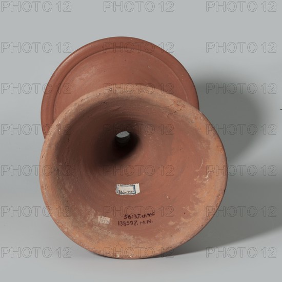 Bowl base. Base for a vessel with almost equal diameter of both rims; both rims irregular with a wavy line. Center of the base is empty. Narrowest part of the base is approximately 2/3 of the height of the entire vessel. Vessel made of Nile clay NB1. Surface leveled, covered with an engobe and polished. Vessel color red. Complete, very well preserved. c. 2494–c. 2181 BC; Old Kingdom, 5th–6th Dynasty, Edfu find place. Vessel; top diameter 17.5 cm, bottom diameter 18 cm, height 25.6 cm, weight 1.30 kg., A single terracotta object with a flared rim and hollow center is shown resting on a smooth light gray surface and casting a soft shadow; the object displays earthy reddish-brown and clay tones with areas of lighter beige wear, a rough textured interior and rim with small imperfections and cracks, and a darker hollow opening at its center, while the background is a uniform pale gray.