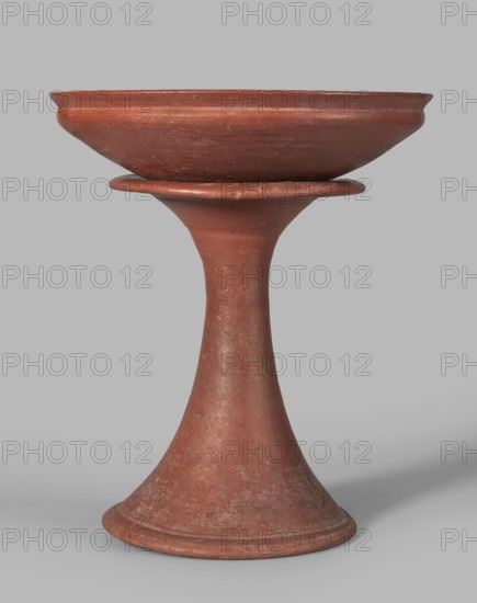 Medum-type bowl, Egyptian pottery, Medum-type bowl, clay vessel. Bowl with a hemispherical shape narrowing just below the rim to form a carination; rounded bottom; straight, slightly widening spout whose diameter is slightly larger than the diameter of the shoulders. Surface even, covered with a red engobe and polished. Vessel color: red. Fairly well preserved with minor losses on the rim. c. 2494–c. 2181 BC; Old Kingdom, 5th–6th Dynasty, Edfu find location. Vessel; height 7 cm, spout diameter 26.4 cm; weight 0.90 kg., A single terracotta object with a reddish-brown color stands centered against a light gray background; it has a wide shallow bowl at the top resting on a flared disk, a tapering, hourglass-like stem, and a broad flared base showing a textured, slightly worn surface with variations of reddish-brown and some paler, dusty patches.