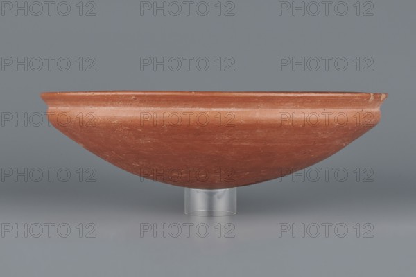 Medum-type bowl, Egyptian pottery, Medum-type bowl, clay vessel. Bowl with a hemispherical shape narrowing just below the rim to form a carination; rounded bottom and straight, slightly widening spout whose diameter is slightly larger than that of the shoulders. Surface even, covered with a red engobe and polished; vessel color red. Fairly well preserved with minor losses on the rim. Circa 2494–2181 BC, Old Kingdom, 5th–6th Dynasty, Edfu. Vessel; height 7 cm, spout diameter 26.4 cm, weight 0.90 kg., A shallow rounded bowl displayed on a short transparent stand, with a smooth matte surface in shades of terracotta and reddish-brown, showing subtle wear marks and lighter scratches; the background and surface beneath are shades of gray and muted bluish-gray.