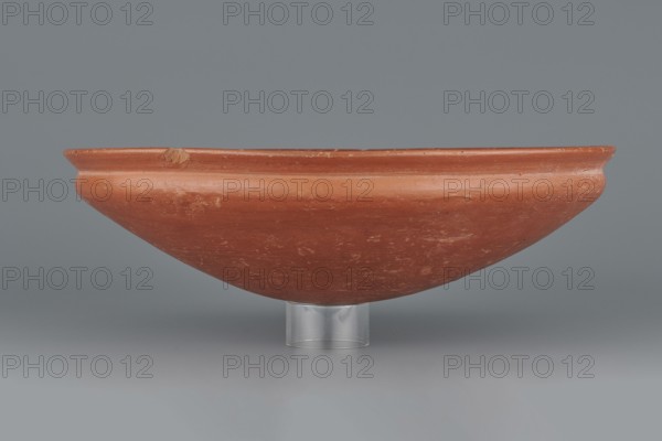 Medum-type bowl, Egyptian pottery, Medum-type bowl, clay vessel with hemispherical shape narrowing just below the rim to form a carination; rounded bottom and a straight, slightly widening spout whose diameter is slightly larger than the shoulders. Surface even, covered with red engobe and polished. Vessel color: red. Fairly well preserved with minor losses on the rim. c. 2494–c. 2181 BC; Old Kingdom, 5th–6th Dynasty, Edfu. Vessel; height 7 cm, diameter 26.4 cm spout, weight 0.90 kg., A shallow, rounded clay bowl with a smooth rim rests on a clear cylindrical stand against a plain background. The bowl is terracotta in color with visible lighter and darker patches and small surface abrasions and scuffs. The rim shows slight irregularities and a small nick. The stand is transparent and colorless, and the background and surface beneath the bowl are shades of gray.