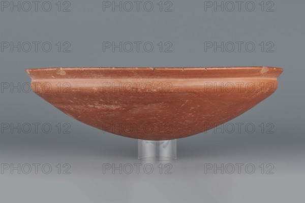 Medum-type bowl, Egyptian pottery, Medum-type bowl, clay vessel. Bowl with a hemispherical shape, narrowing just below the rim to form a carination; rounded bottom and a straight, slightly widening spout whose diameter is slightly larger than that of the shoulders. Surface even, covered with a red engobe and polished. Vessel color: red. Fairly well preserved with minor losses on the rim. Date: c. 2494–c. 2181 BC, Old Kingdom, 5th–6th Dynasty, find location Edfu. Function: vessel. Dimensions: height 7 cm; spout diameter 26.4 cm; weight 0.90 kg., A shallow, broad clay bowl with a rounded underside sits supported by a clear cylindrical stand; the bowl is primarily reddish-brown with lighter worn areas and a few small patches of tan where the surface is chipped, and the background and surface beneath the bowl are shades of gray.