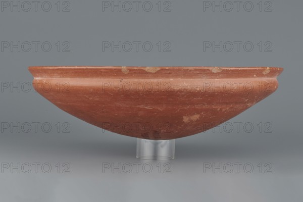 Medum-type bowl, Egyptian pottery, clay vessel. Bowl with a hemispherical shape, narrowing just below the rim to form a carination; rounded bottom; straight, slightly widening spout whose diameter is slightly larger than the shoulders. Surface even, covered with a red engobe and polished. Vessel color: red. Fairly well preserved, minor losses on the rim. Circa 2494–2181 BC; Old Kingdom, 5th–6th Dynasty, Edfu find location. Function: vessel. Height 7 cm; spout diameter 26.4 cm; weight 0.90 kg., A shallow, wide bowl with a rounded profile sits on a short clear cylindrical support against a smooth gray background; the bowl is an earthy reddish-brown color with lighter beige spots and areas of wear, showing surface scratches and subtle variations in tone.