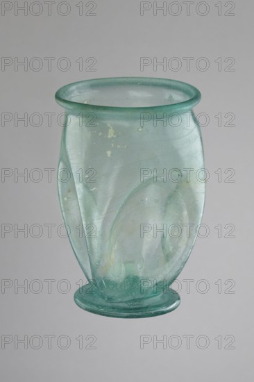 Glass cup, Egypt, 1st–2nd century AD. Type: Isings form 32. Transparent bluish-green glass. Cup with a slightly flaring spout, decorated with four oval concavities on the belly. Ring-shaped foot with a clearance made by pressing the bottom inward; free-blown. Outer diameter of the spout: 6.10 cm. Height: 9 cm. Belly diameter: 6.70 cm. Outer diameter of the bottom: 4.60 cm. Vessel, weight 0.054 kg., A translucent pale green blown-glass vessel with a flared rounded rim and a rounded base on a flat foot, showing a few internal bubbles and subtle surface irregularities, with pale green and faint aqua tones and small areas of light discoloration against a neutral gray background.