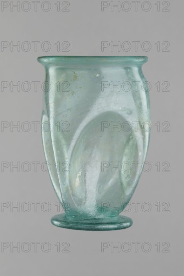 Glass cup, Egypt, 1st–2nd century AD. Isings form 32. Transparent bluish-green glass with slightly flaring spout; decorated with four oval concavities on the belly. Ring-shaped foot with a clearance made by pressing the bottom inward; free-blown. Outer diameter of the spout: 6.10 cm; height: 9 cm; belly diameter: 6.70 cm; outer diameter of the bottom: 4.60 cm; weight 0.054 kg., A translucent glass vessel with a pale green-blue tint is shown against a neutral gray background; the vessel has a rounded rim that flares slightly outward, a bulbous body with smooth, curving folds and subtle surface imperfections, and a flat circular foot; the glass displays areas of cloudiness, small bubbles, and a few pale deposits, with visible variations in green, blue, teal, and clear tones across its surface.