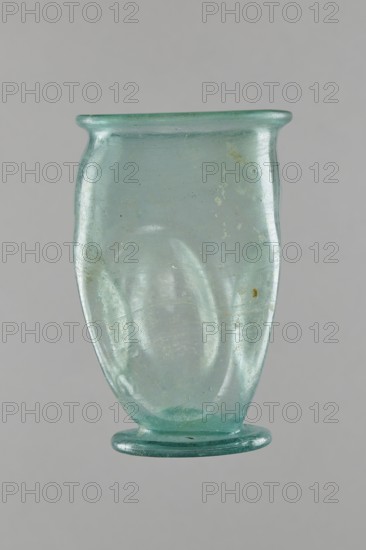 Glass cup, Egypt, 1st–2nd century AD. Isings form 32. Transparent bluish-green glass with slightly flaring spout; decorated with four oval concavities on the belly. Ring-shaped foot with a clearance made by pressing the bottom inward; free-blown. Outer diameter of spout: 6.10 cm; height: 9 cm; belly diameter: 6.70 cm; outer diameter of bottom: 4.60 cm. Vessel; weight 0.054 kg., A translucent pale green, slightly bluish glass vessel with a flared rim and a rounded body that tapers inward toward a flat, circular foot; the surface shows irregularities and small bubbles, with areas of faint cloudiness and a few pale discolorations, all photographed against a neutral light gray background.