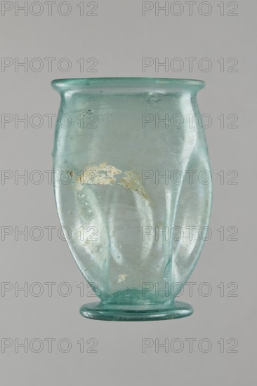 Glass cup, Egypt, 1st–2nd century AD. Type: Isings form 32. Transparent bluish-green glass. Cup with a slightly flaring spout, decorated with four oval concavities on the belly. Ring-shaped foot with a clearance made by pressing the bottom inward; free blown. Outer diameter of the spout: 6.10 cm. Height: 9 cm. Belly diameter: 6.70 cm. Outer diameter of the bottom: 4.60 cm. Vessel, weight 0.054 kg., A translucent pale green glass vessel with a flared rim and a rounded body tapering to a flattened foot, showing surface irregularities and a few rough patches of encrustation, with subtle internal striations and small air bubbles visible in the glass against a neutral gray background.