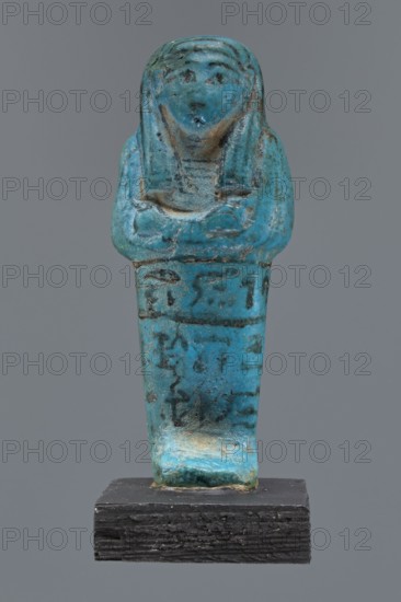 Ushabti Cha-em-uase. A mummy-shaped figurine shabti made of faience and pressed into a mold. Details of the face, jewelry, and bag are painted. Hands folded across the chest—right over left. Wide bracelets on the wrists. An ush necklace on the chest. No tools in the hands; a bag on the back. A three-part wig, combed into strips, with borders at the ends. Painted inscription, three lines of text on the legs: Shd Osiris Cha-em-Wese justified with his voice said: O — the beginning of the sixth chapter of the Book of the Dead. Ca. 1295–1069 BC; New Kingdom, 19th–20th Dynasty. Height 10.1 cm, width 4.1 cm. Maker, private name Cha-em-uase., A small statuette shaped like a standing human figure with folded arms, rendered in a bright turquoise-blue material with areas of darker blue and greenish patina and some brownish and blackened spots, mounted on a dark textured rectangular base; the figure has a rounded head with stylized facial features and a headdress-like form extending down the sides...
