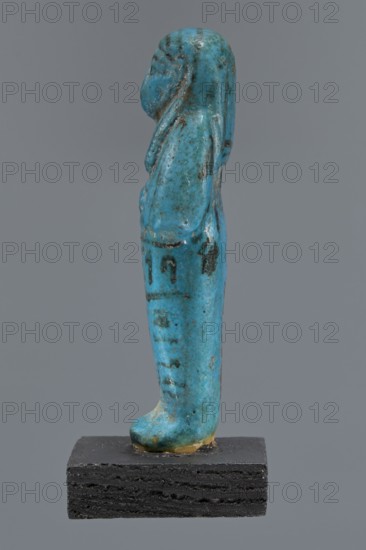 Ushabti Cha-em-uase, Cha-em-uase private name. A mummy-shaped shabti figurine made of faience, pressed in a mold; details of the face, jewelry, and bag are painted. Hands folded across the chest right over left. Wide bracelets on the wrists and an ush necklace on the chest. No tools in the hands; a bag on the back. Wears a three-part wig combed into strips with borders at the ends. Painted inscription of three lines on the legs: Shd Osiris Cha-em-Wese justified with his voice said: O — the beginning of the sixth chapter of the Book of the Dead. New Kingdom, 19th–20th Dynasty, ca. 1295–1069 BC. Shabti; height 10.1 cm, width 4.1 cm. Typology according to Schneider. Comparative material: H. Ranke, Die Agyptischen Personennamen I, 1935, p. 263,19., A small upright figurine in a bright turquoise blue with darker blue-green speckles and some brownish-black spots stands on a rectangular black base; the figure has a rounded head with a hint of facial profile and a long headdress or hair that d...