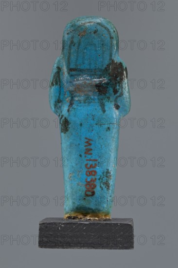 Ushabti Cha-em-uase, Cha-em-uase private name. A mummy-shaped figurine shabti made of faience, pressed into a mold. Details of the face, jewelry, and bag are painted. Hands folded across the chest—right over left. Wide bracelets on the wrists. An ush necklace on the chest. No tools in the hands; a bag on the back. A three-part wig, combed into strips, with borders at the ends. Painted inscription, three lines of text on the legs: Shd Osiris Cha-em-Wese justified with his voice said: O — the beginning of the sixth chapter of the Book of the Dead. Ca. 1295–1069 BC; New Kingdom, 19th–20th Dynasty, Egypt. Height 10.1 cm, width 4.1 cm., A small figurine shaped like a standing wrapped figure with a rounded head and simplified arms, displayed on a rectangular dark base, predominantly bright turquoise blue with areas of darker blue and black discoloration and small spots of brownish wear, and a thin band of yellowish residue along the bottom edge where it meets the base.