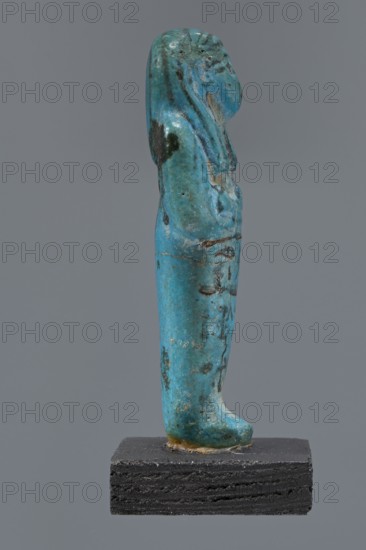 Ushabti Cha-em-uase, Cha-em-uase private name. A mummy-shaped figurine ushabti made of faience and pressed into a mold. Details of the face, jewelry, and bag are painted. Hands folded across the chest—right over left. Wide bracelets on the wrists. An ush necklace on the chest. No tools in the hands, a bag on the back. A three-part wig, combed into strips, with borders at the ends. Painted inscription of three lines placed on the legs: Shd Osiris Cha-em-Wese justified with his voice said: O — the beginning of the sixth chapter of the Book of the Dead. ca. 1295–1069 BC; New Kingdom, 19th–20th Dynasty, Egypt. Height 10.1 cm, width 4.1 cm., A small standing figurine displayed on a rectangular dark base, the figurine rendered in a turquoise blue material with areas of darker blue and greenish tones and some black and brown spots and traces; the figure is shown in profile with a smooth, elongated body, a rounded head with stylized hair or headdress that falls along the back, a simple facial ...