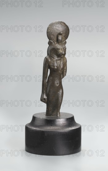 Sekhmet figurine, Figurine of the lion-headed goddess Sekhmet, shown standing or walking. The figure has a lioness head in a tripartite wig topped with a solar disk adorned with a uraeus. Sekhmet's right arm hangs down at her right side, clenched into a fist, showing a hole left by a lost cult object; the left arm was broken at the elbow. The figurine was broken at Sekhmet's knuckles and is mounted on a modern cylindrical wooden base with a contoured edge. 664–525 BC; Late Period, 26th Dynasty, Egypt. Figurine, height 9.7 cm., A small bronze statuette of a standing anthropomorphic figure with an animal-like head is shown mounted on a round dark pedestal; the figure has a slender elongated body, one arm hanging at the side and the other bent behind the back, and a circular disk behind the head. The visible colors are dark brown and bronze tones on the statuette and black on the pedestal, set against a light gray background.