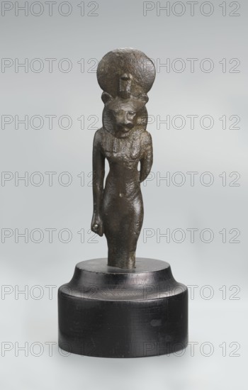Sekhmet figurine, lion-headed goddess shown standing or walking. The figure has a lioness' head in a tripartite wig topped with a solar disk adorned with a uraeus. Sekhmet's right arm hangs down at her side, clenched into a fist, with a hole left by a lost cult object; the left arm is broken at the elbow. The figurine was broken at the knuckles and is mounted on a modern cylindrical wooden base with a contoured edge. 664–525 BC; Late Period, 26th Dynasty, Egypt. Terracotta/stone figurine, height 9.7 cm., A small sculpted figure stands on a rounded dark base against a pale gray background; the figure is dark bronze with a textured surface and depicts a slender humanlike body with a feline head topped by a circular disk and a small vertical projection at the disk's front, the head showing carved facial features and rounded ears, the torso showing faint indications of a collar or necklace and one arm hanging down while the other is bent behind the back, and the base is a smooth black or v...