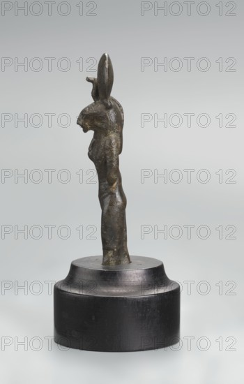Sekhmet figurine, lion-headed goddess shown standing or walking. Lioness head with a tripartite wig topped by a solar disk adorned with a uraeus. Right arm hangs at her side, clenched into a fist, revealing a hole from a lost cult object; left arm broken at the elbow. Figurine broken at the knuckles and mounted on a modern cylindrical wooden base with a contoured edge. 664–525 BC; Late Period, 26th Dynasty, Egypt. Terracotta/stone figurine, height 9.7 cm., A small three-dimensional metal figure with a dark brown and greenish patina stands upright on a round black base; the figure is elongated and stylized with an upright posture, a pronounced head with an elongated vertical element rising above it, a slightly protruding snout or muzzle, and a tapered body that narrows toward the waist and hips, showing subtle surface irregularities and textural variations across the metal.