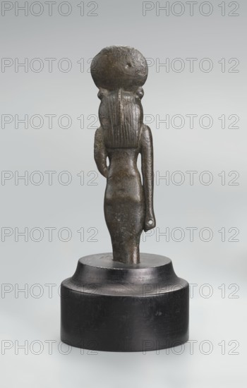 Sekhmet figurine, lion-headed goddess shown standing or walking; lioness head with tripartite wig topped by a solar disk adorned with a uraeus. Right arm hangs at her side, clenched into a fist revealing a hole from a lost cult object; left arm broken at the elbow. Figurine broken at the knuckles and mounted on a modern cylindrical wooden base with a contoured edge. 664–525 BC; Late Period, 26th Dynasty, Egypt; figurine; height 9.7 cm., A small dark bronze statue of a standing human figure is shown from the back, placed on a round dark wooden base; the figure has a smooth rounded head with a large circular disk on top, long straight hair carved in vertical grooves, a narrow waist and hips, and one arm bent with the hand resting at the side while the other arm hangs down with a small hole near the wrist; the statue surface shows a mottled dark brown and greenish patina with some lighter worn spots, and the base is a deep black with a slightly glossy finish, all set against a soft pale g...