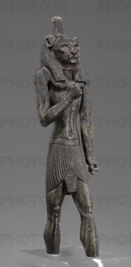 Figurine of the god Mahes. Figurine of a striding man with a lion's head. The lion's head is set directly on the shoulders. The wig is striped. A square pin is on top of the head. The right arm is bent at the elbow and placed on the chest, the left arm is freely placed along the body. A short striped loincloth is on the hips., Mahes myth.. Egyptian deities; Egyptian art; Egyptian sculpture. Late Period, 26th Dynasty, 664–525 BC, Egypt. Figurine; height 26 cm, width 7 cm, depth 3.8 cm., A dark gray statuette of a standing anthropomorphic figure with a feline head and a tall cylindrical crown, showing a textured mane and carved facial features, wearing a pleated skirt with detailed vertical and diagonal patterns and a narrow decorative belt, with one arm bent across the chest and the other hanging down holding a small rounded object, the surface showing areas of lighter wear and small patches of brownish discoloration, set against a uniform medium gray background and mounted on a clear b...