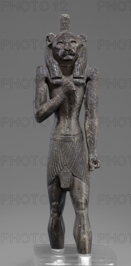 Figurine of the god Mahes. Mahes myth.. Egyptian deities, Egyptian art, Egyptian sculpture. Figurine of a striding man with a lion's head. The lion's head is set directly on the shoulders. The wig is striped. A square pin is on top of the head. The right arm is bent at the elbow and placed on the chest, the left arm is freely placed along the body. A short striped loincloth is on the hips. 664–525 BC; Late Period, 26th Dynasty, Egypt. Figurine; height 26 cm, width 7 cm, depth 3.8 cm., A dark gray-green metal statue of a standing anthropomorphic figure with a feline head and mane, wearing a patterned skirt and a short apron-like panel, with one arm bent across the chest and the other hanging at the side, showing textured surface wear and small lighter patches of corrosion, set against a plain neutral gray background and mounted on a clear rectangular base.