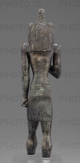 Figurine of the god Mahes. Mahes myth., Egyptian deities, Egyptian art, Egyptian sculpture. Figurine of a striding man with a lion's head. The lion's head is set directly on the shoulders. The wig is striped. A square pin is on top of the head. The right arm is bent at the elbow and placed on the chest, the left arm is freely placed along the body. A short striped loincloth is on the hips. 664–525 BC; Late Period, 26th Dynasty, Egypt. Figurine; height 26 cm, width 7 cm, depth 3.8 cm., A weathered dark bronze figurine viewed from the back stands on a clear base against a neutral gray background; the figure has a rounded headpiece with carved vertical lines and a central vertical protrusion, small rounded ear-like shapes, broad shoulders and muscular arms with one arm bent and the other hanging down holding a small object, a narrow waist leading to a pleated skirt with textured vertical ridges and a patterned belt area, and short sturdy legs showing patches of lighter greenish corrosion ...