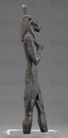 Figurine of the god Mahes, Mahes myth., Egyptian deities, Egyptian art, Egyptian sculpture. Figurine of a striding man with a lion's head. The lion's head is set directly on the shoulders. The wig is striped. A square pin is on top of the head. The right arm is bent at the elbow and placed on the chest, the left arm is freely placed along the body. A short striped loincloth is on the hips. 664–525 BC; Late Period, 26th Dynasty, Egypt. Figurine; height 26 cm, width 7 cm, depth 3.8 cm., A dark gray metal statue of a standing anthropomorphic figure with a feline head, wearing a long textured headdress and a pleated skirt, shown in profile against a neutral gray background; the figure's arms are held bent at the chest with hands clasped, the surface shows rough patina with hints of lighter gray and subtle greenish corrosion, and it stands on a transparent base with reflections.