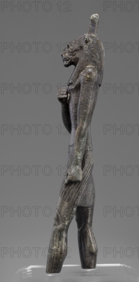 Figurine of the god Mahes, Mahes myth., Egyptian deities, Egyptian art, Egyptian sculpture. Figurine of a striding man with a lion's head. The lion's head is set directly on the shoulders. The wig is striped. A square pin is on top of the head. The right arm is bent at the elbow and placed on the chest, the left arm is freely placed along the body. A short striped loincloth is on the hips. 664–525 BC; Late Period, 26th Dynasty, Egypt. Figurine; height 26 cm, width 7 cm, depth 3.8 cm., A standing statue of a humanoid figure with an animal-like head is shown in profile against a plain gray background, rendered in dark metallic tones with patches of greenish and brownish patina; the figure wears a fitted skirt with textured vertical patterns and a narrow collar, has a tall headdress extending above the head, and one arm folded across the chest while the other hangs by the side, with visible surface roughness and weathered areas on the torso, limbs, and head.