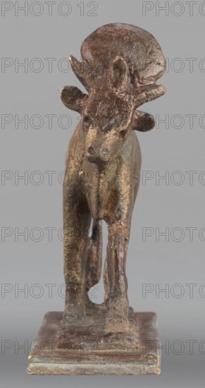 Apis bull figurine, Apis mythology, bulls, sacred animals, animal figurines, Egyptian art, Sacred animal of the god Ptah, 664–525 BC; Late Period, 26th Dynasty, Egypt, figurine, height 7 cm, width 6 cm, weight 0.164 kg, A small sculptural object depicting an animal standing on a rectangular base, rendered in a metallic material with a rough, textured surface; the figure shows a head with protrusions and rounded ears, a tapered snout, a torso with visible limbs positioned on the base, and an uneven, pitted patina. Colors visible include various shades of brown, bronze, and gold with hints of greenish oxidation and darker brown areas, set against a plain light gray background.